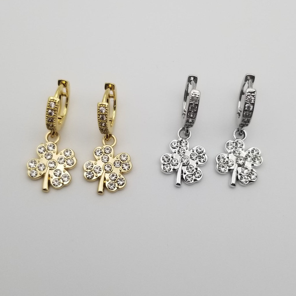Two pairs of clover-shaped earrings with cubic zirconia, one gold and one silver, on a light gray background.