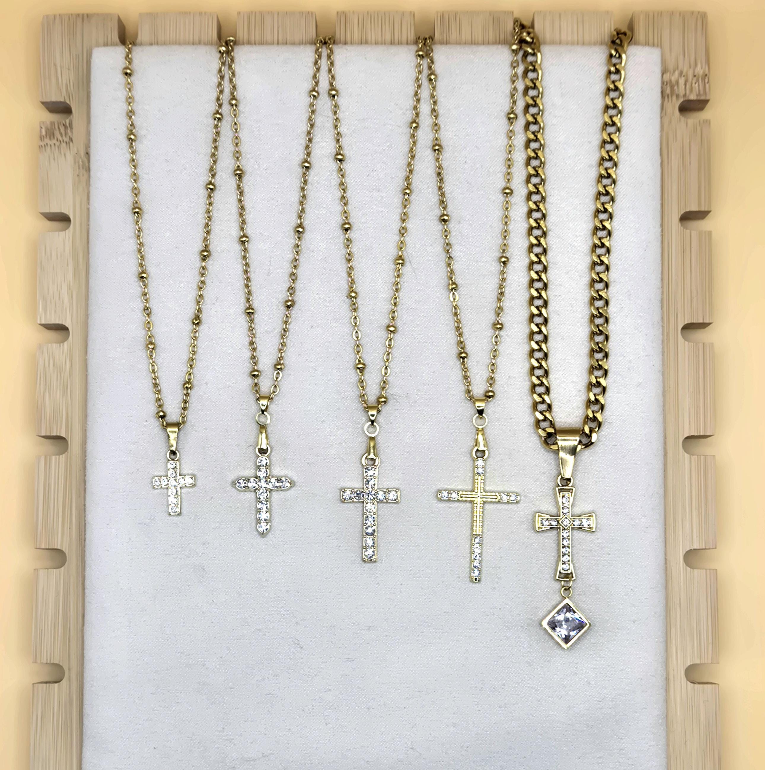 A display of five 18k gold-plated cross necklaces on a white jewelry board. Four pendants feature shimmering cubic zirconia on dainty satellite chains, while the fifth is an ornate cross with a square crystal drop on a bold Cuban link chain. Elegant Christian jewelry for layering or daily wear.