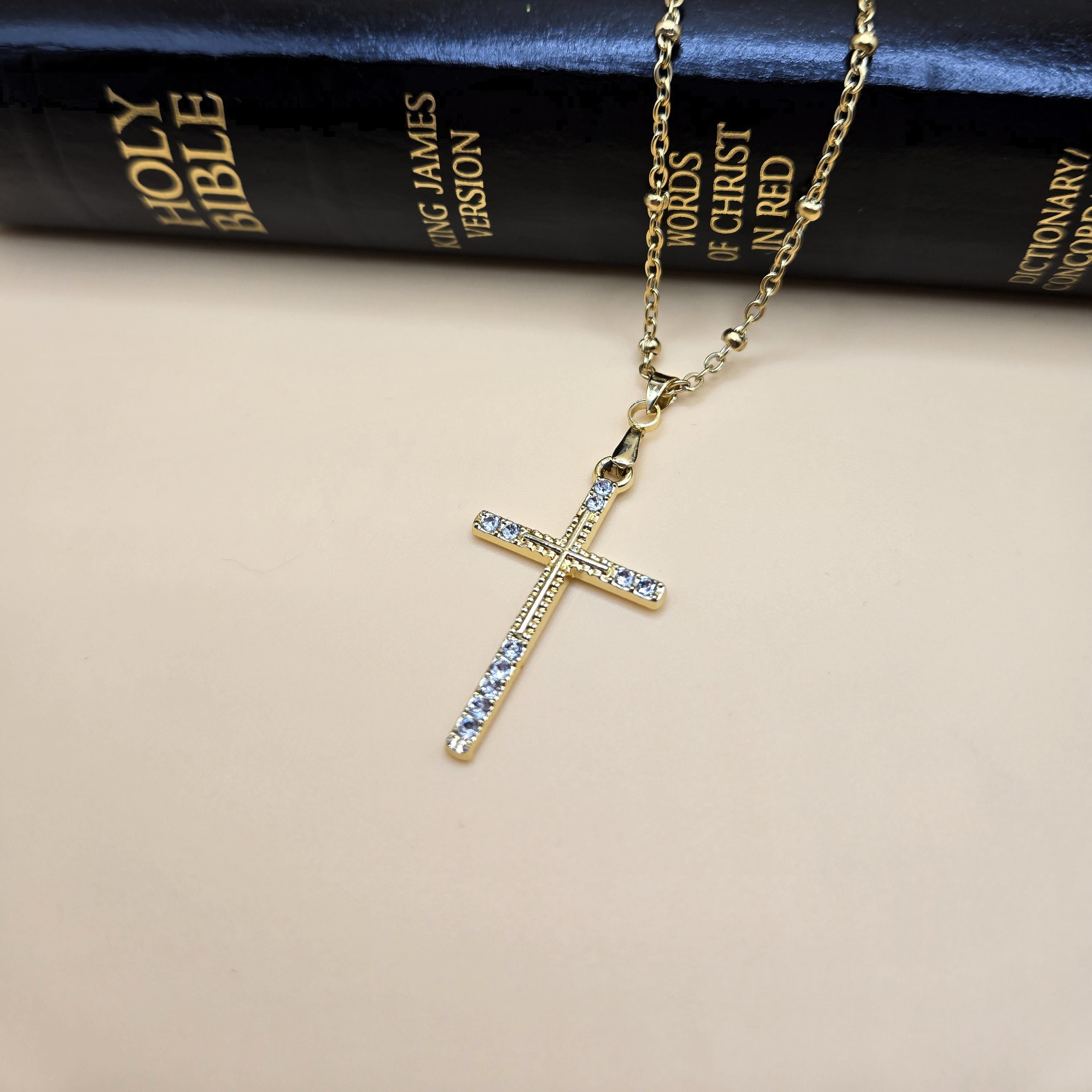 A close-up of a slender 18k gold-plated cross necklace accented with cubic zirconia on a delicate satellite chain. The jewelry is elegantly draped over a black King James Version Holy Bible, emphasizing the connection between faith and quality craftsmanship.