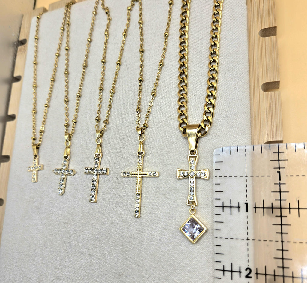 A size comparison of five 18k gold-plated cross necklaces with cubic zirconia on a jewelry display. A ruler is placed next to the largest ornate cross pendant to show it measures approximately 1.5 inches in length. The image highlights the different pendant sizes and chain styles, including dainty satellite and bold Cuban link.