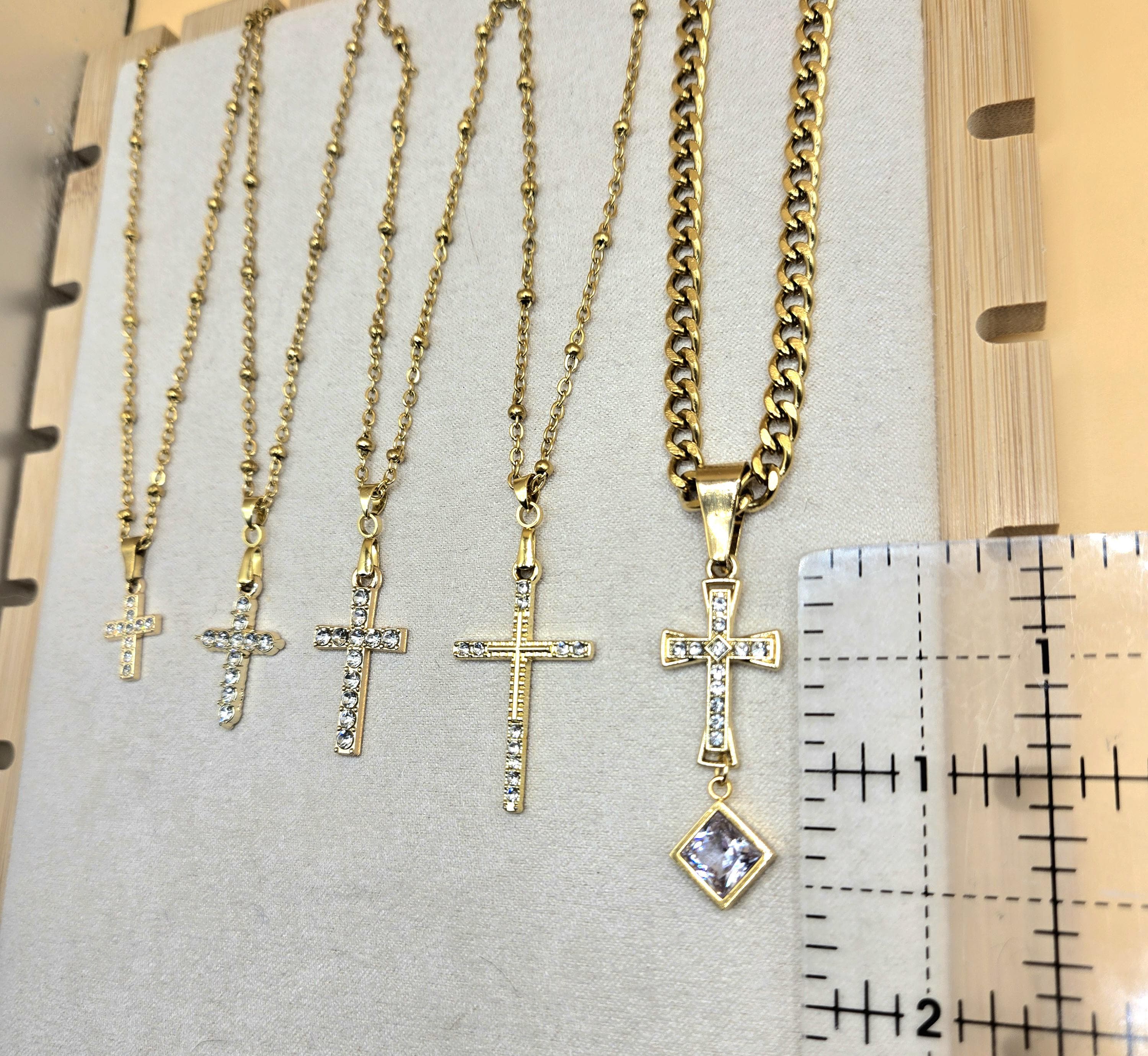 A size comparison of five 18k gold-plated cross necklaces with cubic zirconia on a jewelry display. A ruler is placed next to the largest ornate cross pendant to show it measures approximately 1.5 inches in length. The image highlights the different pendant sizes and chain styles, including dainty satellite and bold Cuban link.