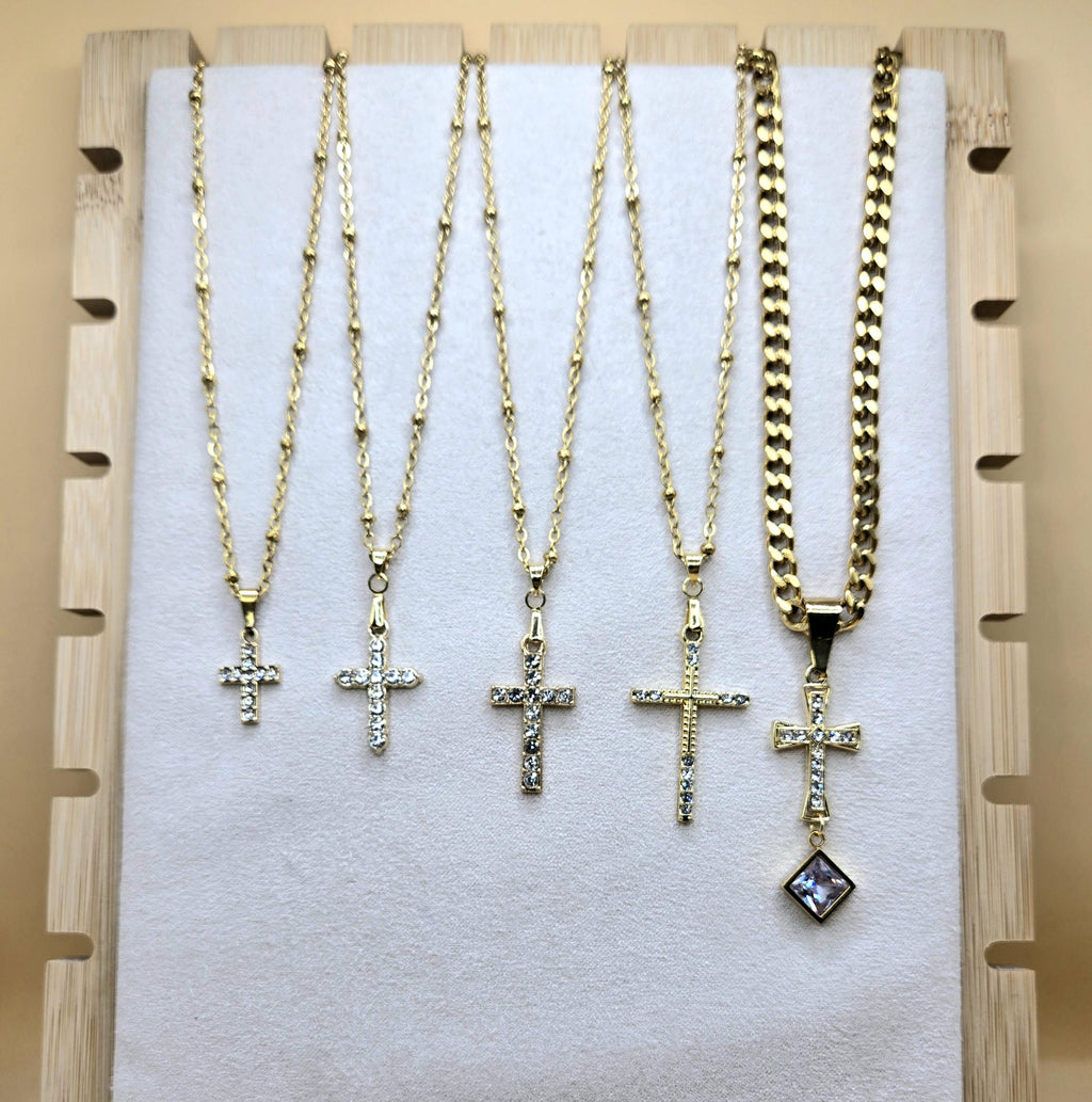A display of five 18k gold-plated cross necklaces on a white jewelry board. Four pendants feature shimmering cubic zirconia on dainty satellite chains, while the fifth is an ornate cross with a square crystal drop on a bold Cuban link chain. Elegant Christian jewelry for layering or daily wear.
