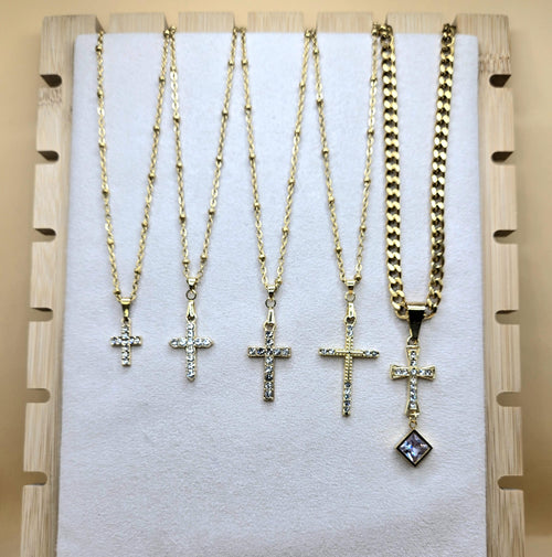 A display of five 18k gold-plated cross necklaces on a white jewelry board. Four pendants feature shimmering cubic zirconia on dainty satellite chains, while the fifth is an ornate cross with a square crystal drop on a bold Cuban link chain. Elegant Christian jewelry for layering or daily wear.