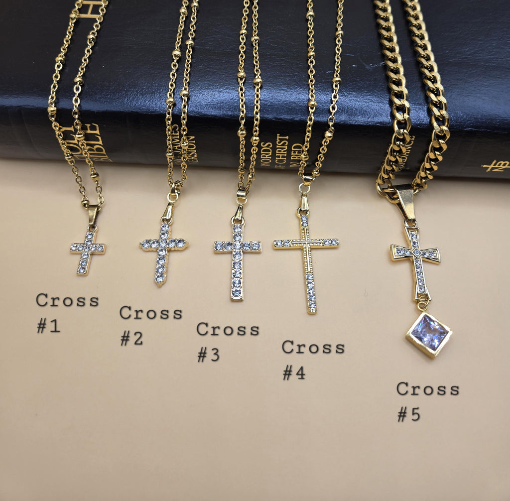 A collection of five unique 18k gold-plated cross necklaces with cubic zirconia crystals, labeled Cross #1 through Cross #5. The display features minimalist and ornate designs on satellite and cuban link chains, presented over a King James Version Bible as a symbol of faith and the Word of God.