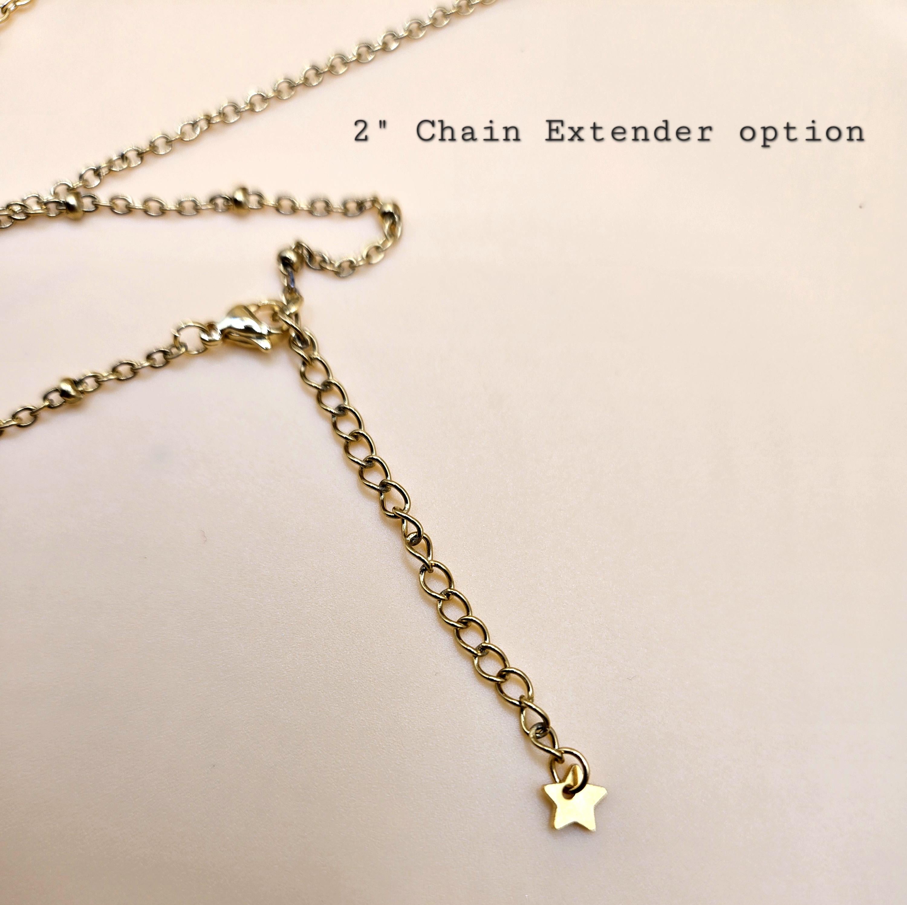 A close-up view of the 2-inch 18k gold-plated chain extender option. The extension chain features a tiny gold star charm at the end and is shown attached to a lobster claw clasp, demonstrating the adjustable fit available for layering these cross necklaces.