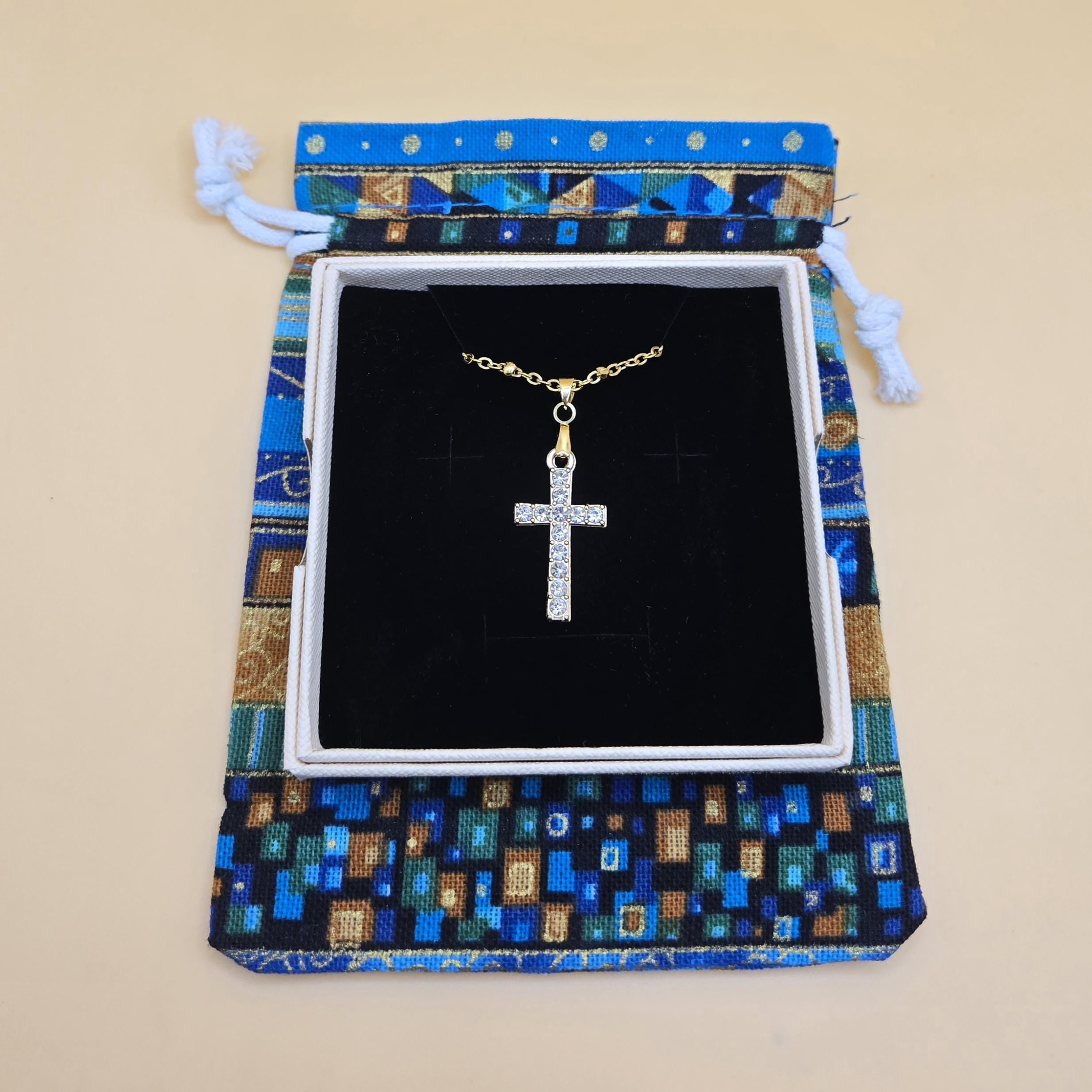 An 18k gold-plated cross necklace with cubic zirconia crystals, nestled in a white jewelry gift box. The box is resting on a colorful, mosaic-patterned blue and gold drawstring pouch, showcasing the elegant gift-ready packaging.
